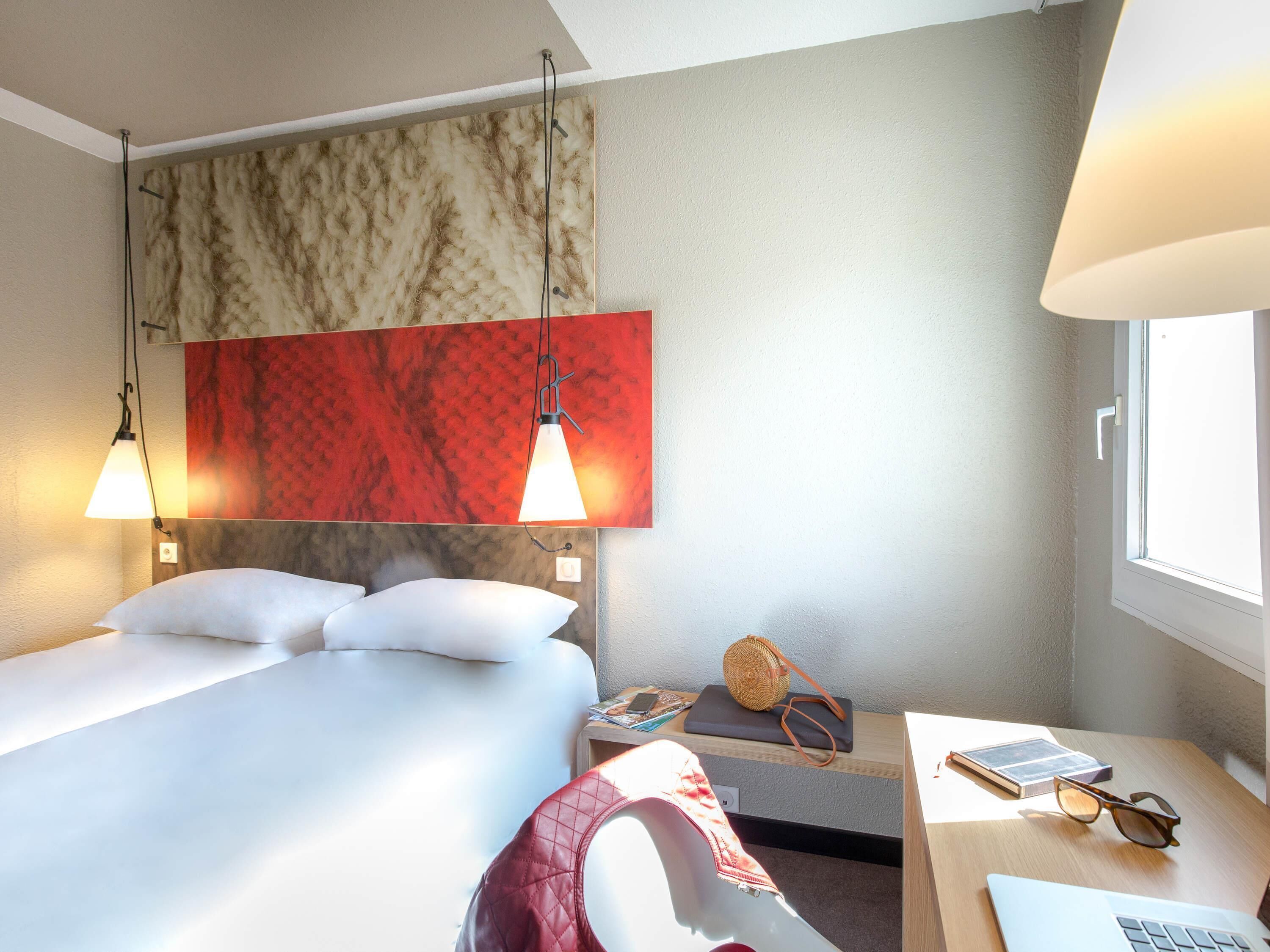Habitación de hotel con una cama, dos lámparas colgantes, una mesita de noche con una computadora portátil, gafas y una revista.
