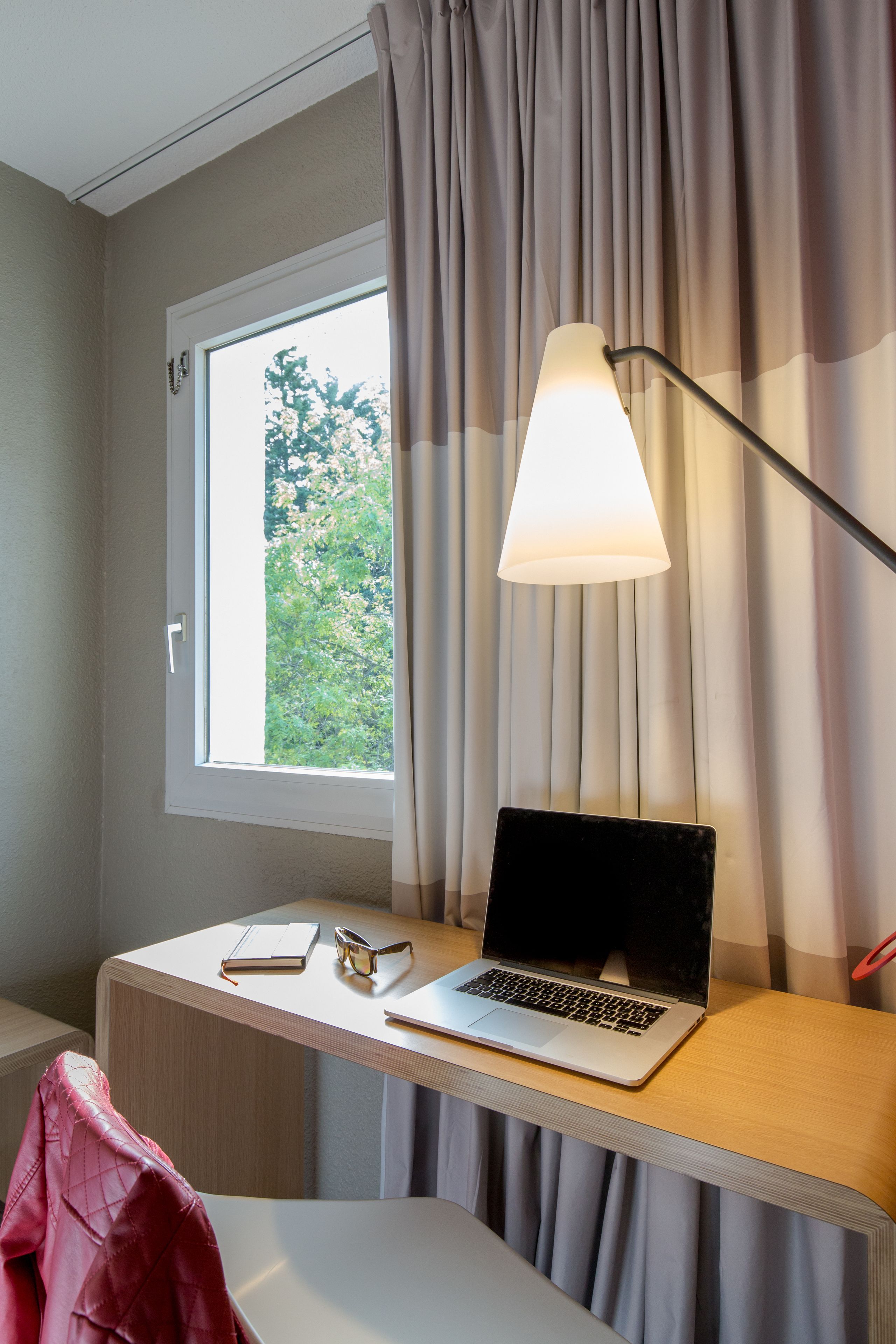 Una habitación con escritorio, laptop, lámpara y ventana con cortinas.