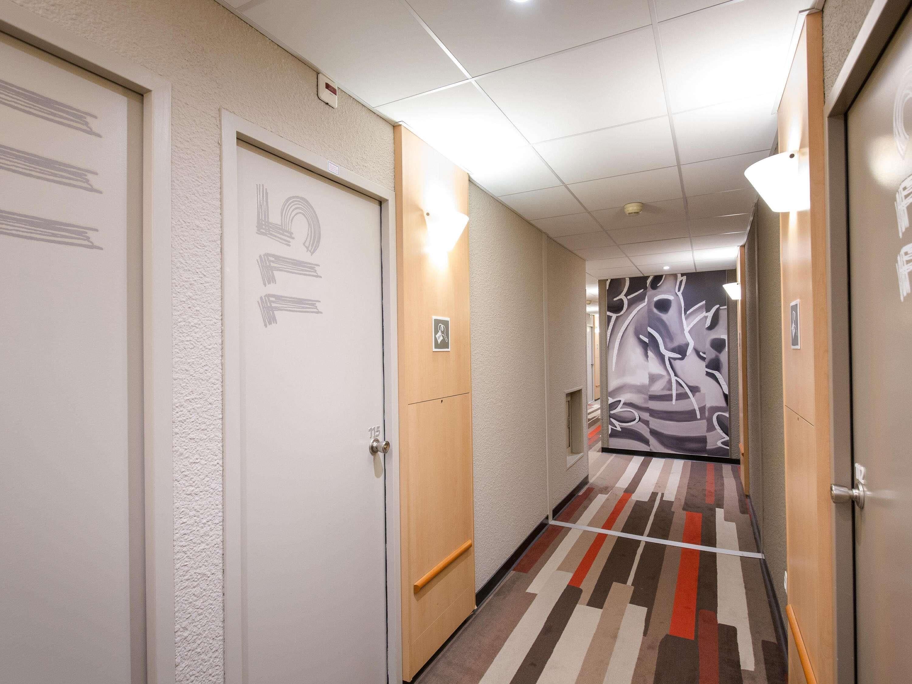 A hotel hallway with doors numbered 115, a patterned wall mural, and a striped carpet.
