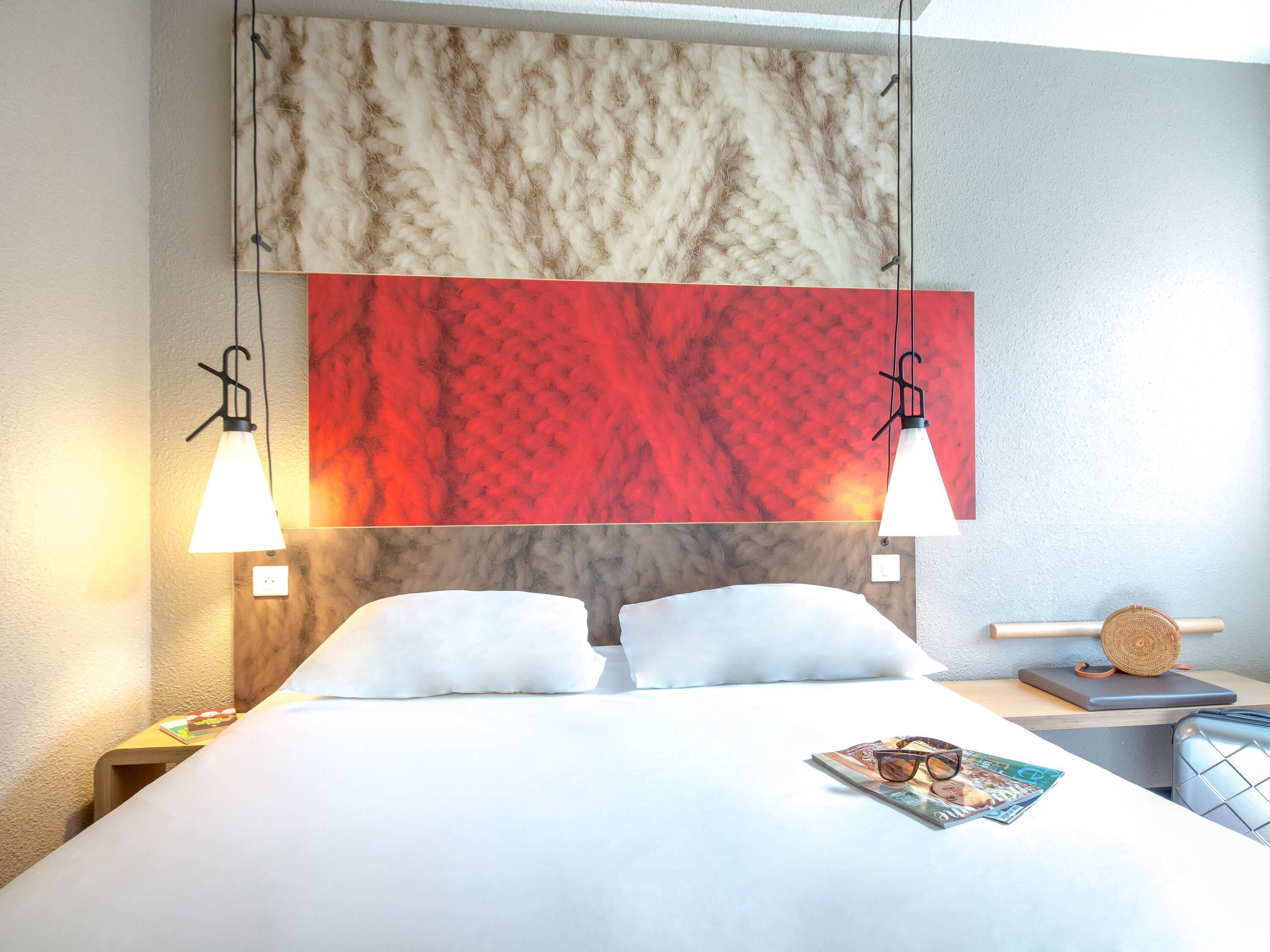 A hotel room with a bed, two bedside lamps, a magazine on the bed, and a small table with a basket.