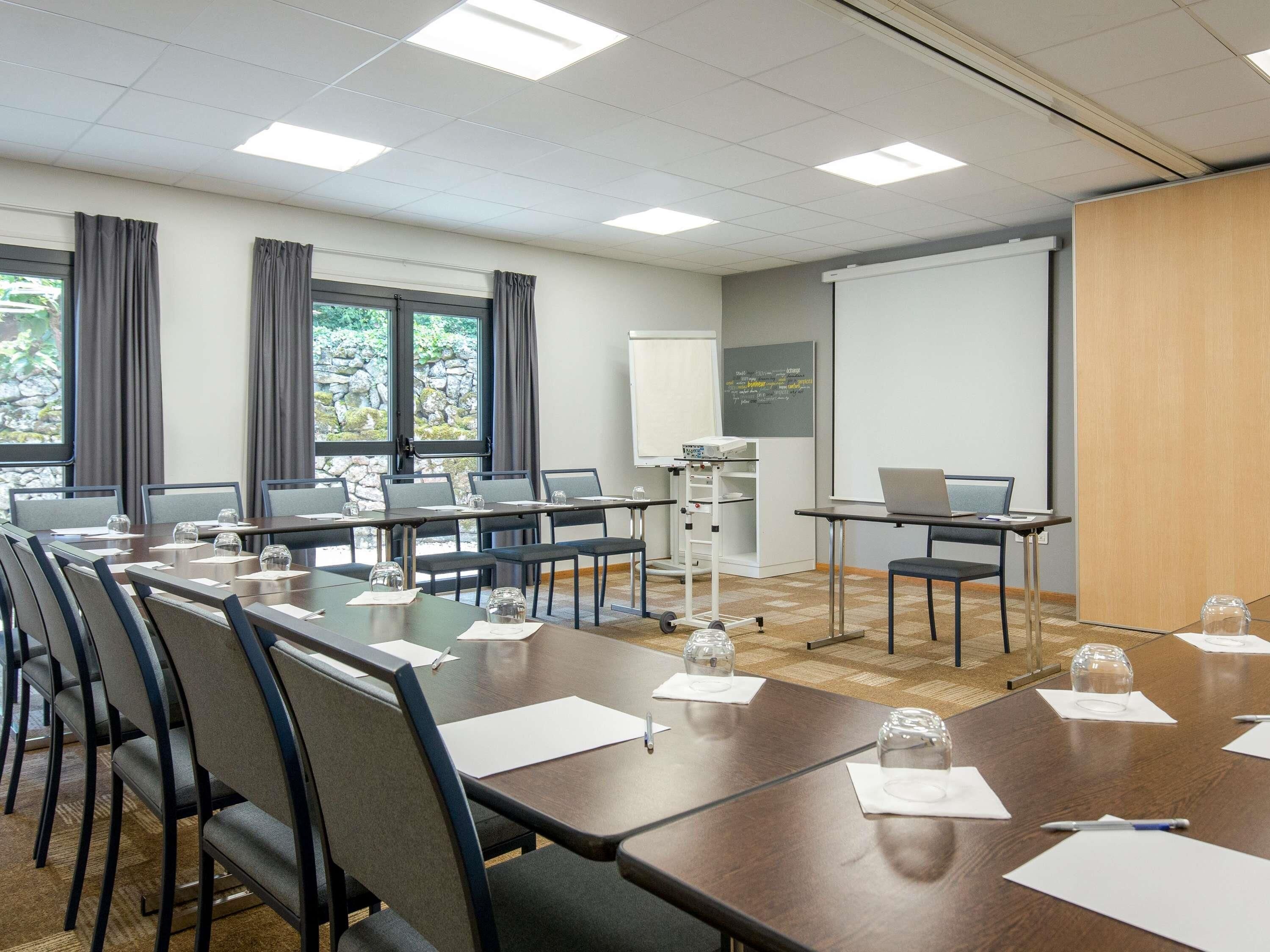 Une salle de conférence avec une longue table, des chaises, un écran de projection et un ordinateur portable posé sur un bureau.