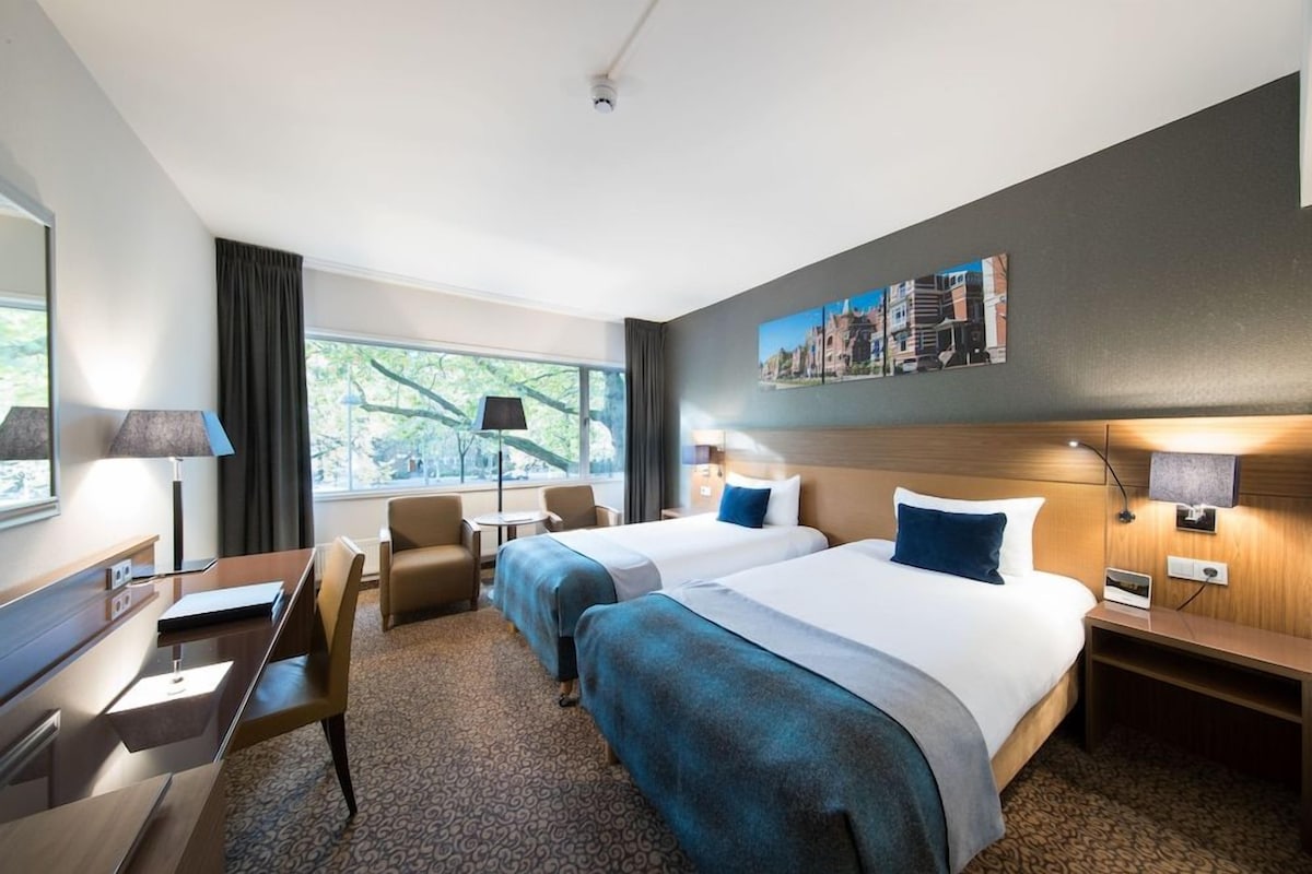 A hotel room with two beds, a desk, a chair, a small table, and a window with curtains.