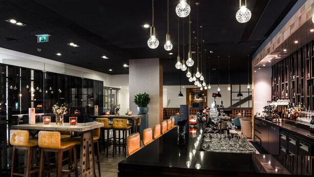 A modern bar area with a long counter, multiple stools, hanging pendant lights, and a bar stocked with bottles. - Bilderberg Garden Hotel (Amsterdam)