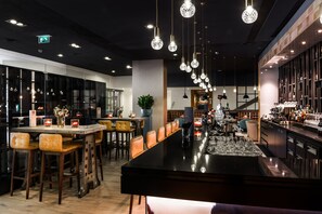A modern bar area with a long counter, multiple stools, hanging pendant lights, and a bar stocked with bottles. - Bilderberg Garden Hotel (Amsterdam)