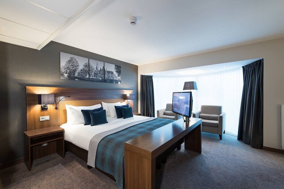 A hotel room with a large bed, a desk, a TV, a chair, and a window with curtains.