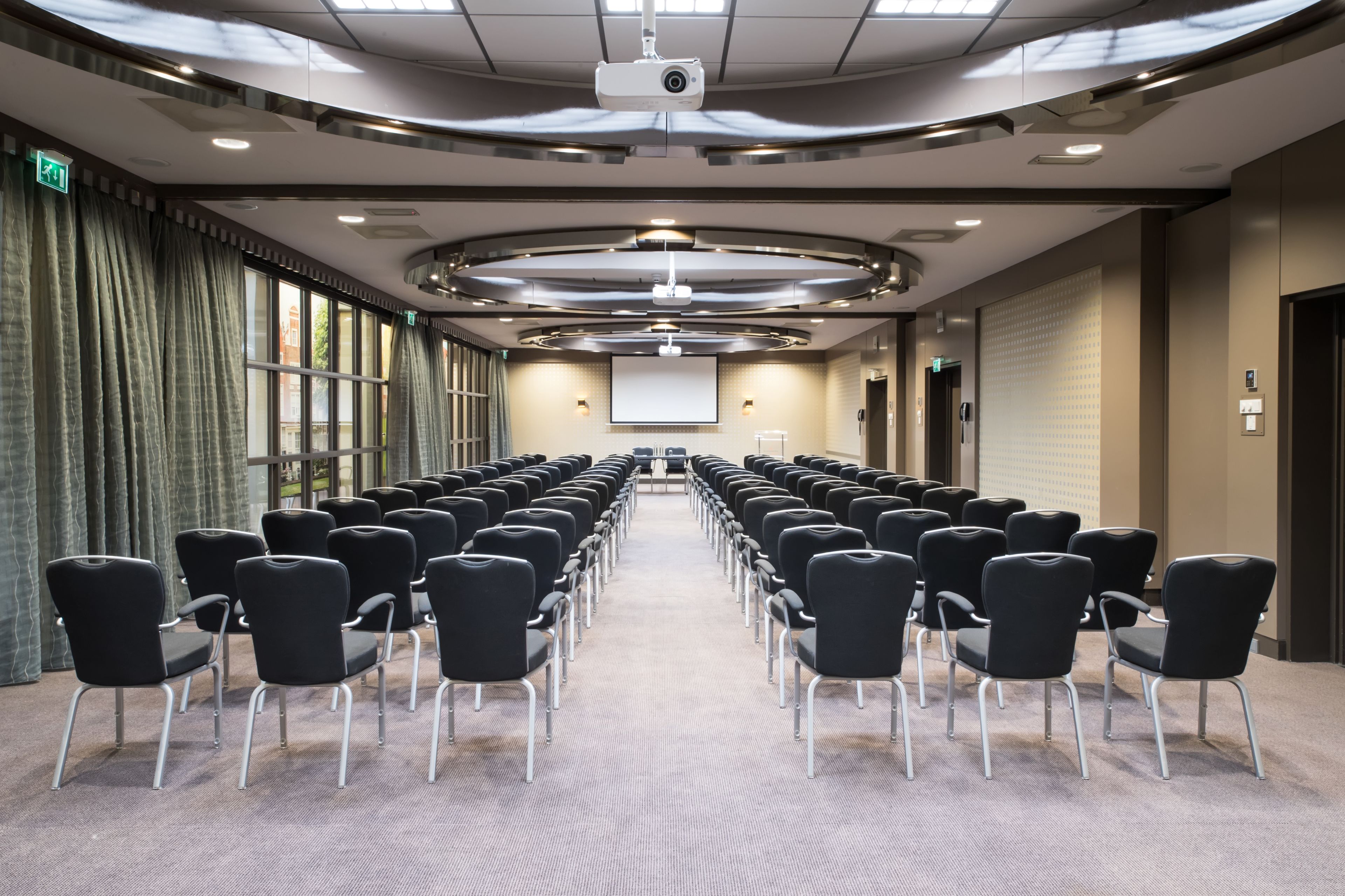 A conference room with rows of black chairs, a projector screen, and a large window with curtains.