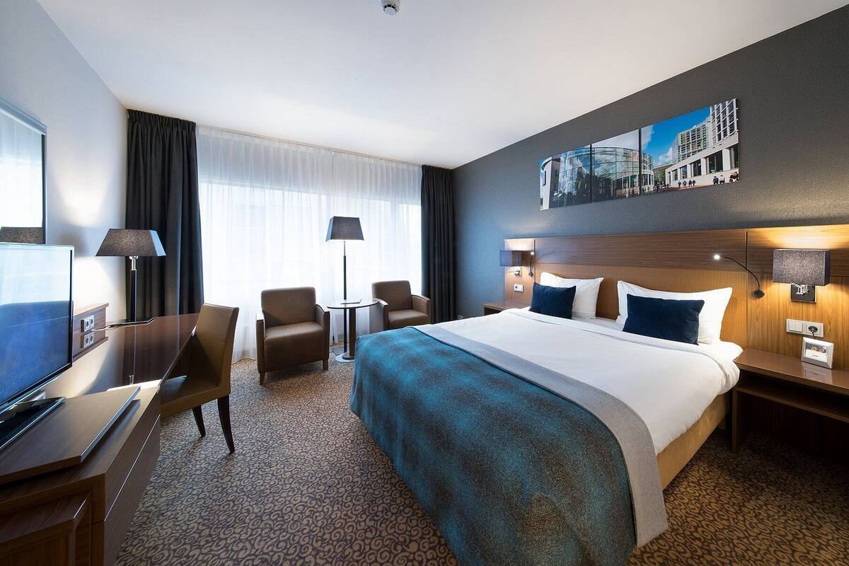 A hotel room with a large bed, a desk with a TV, two armchairs, a lamp, and a painting on the wall.