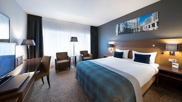 Ein Hotelzimmer mit einem großen Bett, einem Schreibtisch mit Fernseher, zwei Sesseln, einer Lampe und einem Bild an der Wand.