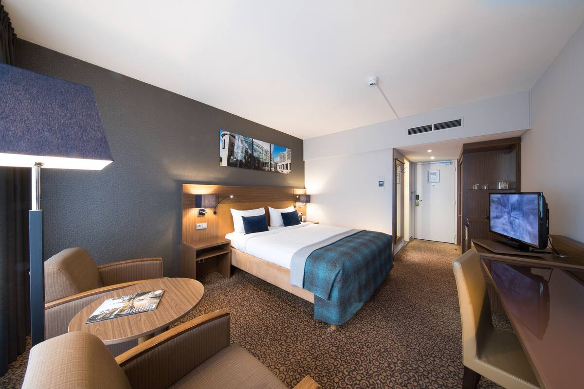 A hotel room with a bed, a desk with a television, a chair, a table, and a lamp.