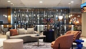 A modern living room with a sectional sofa, round ottoman, and two armchairs. A coffee table is in the center, and the room features a decorative wall with shelves and hanging lights. - Bilderberg Garden Hotel (Amsterdam)