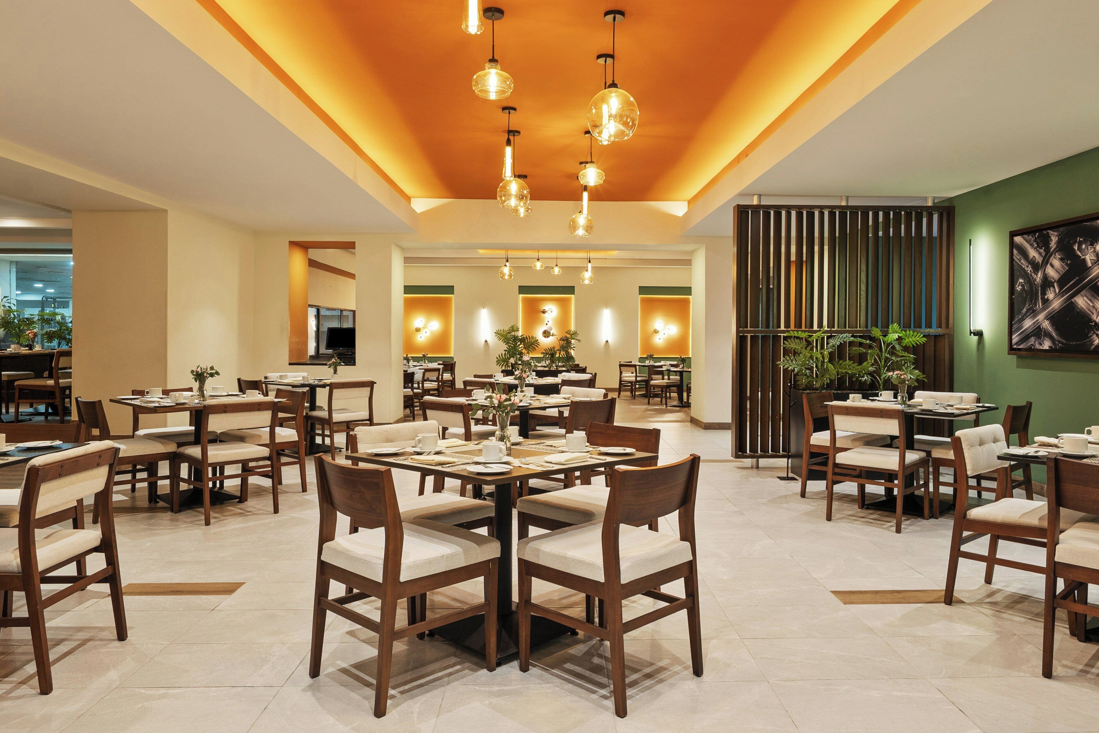 A spacious dining area with multiple tables and chairs, a modern ceiling design with hanging lights, and a view of the kitchen in the background.