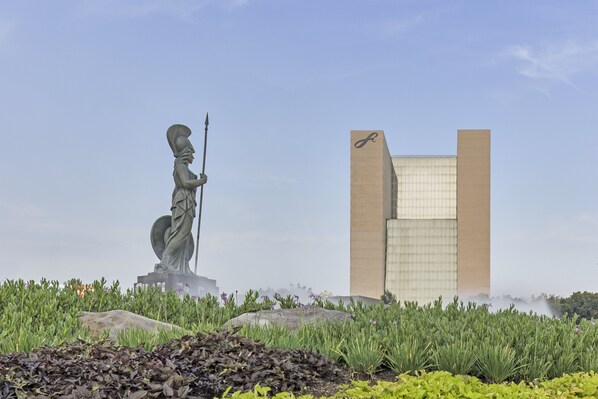 Stunning statue of a figure holding a spear stands amidst lush greenery and a unique rock formation, set against the backdrop of a striking modern building with a grid-like facade.
