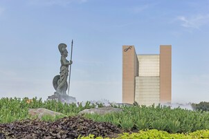 Una estatua de una figura sosteniendo una lanza, un edificio moderno con un logotipo distintivo, y un jardín con diversas plantas y rocas.