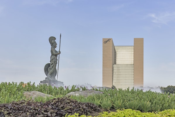 Stunning statue of a figure holding a spear stands amidst lush greenery and a unique rock formation, set against the backdrop of a striking modern building with a grid-like facade.