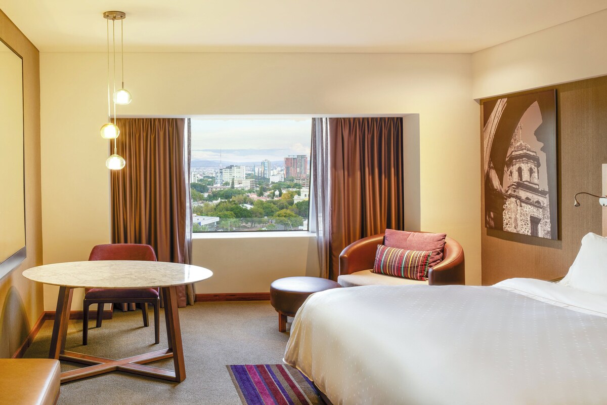 A hotel room with a bed, a desk with a chair, a small table, a TV, and a window with a city view.