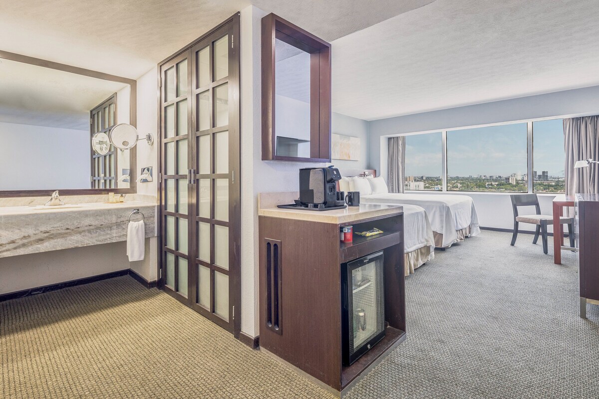 A hotel room with a large window, a minibar, a desk, and a separate bathroom area.