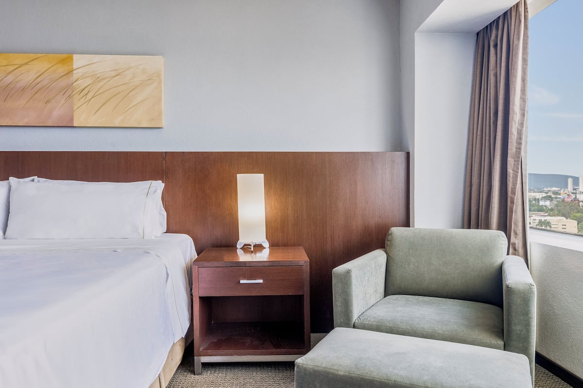 Een hotelkamer met een bed, een nachtkastje met een lamp, een stoel, en een groot raam met uitzicht op de stad.