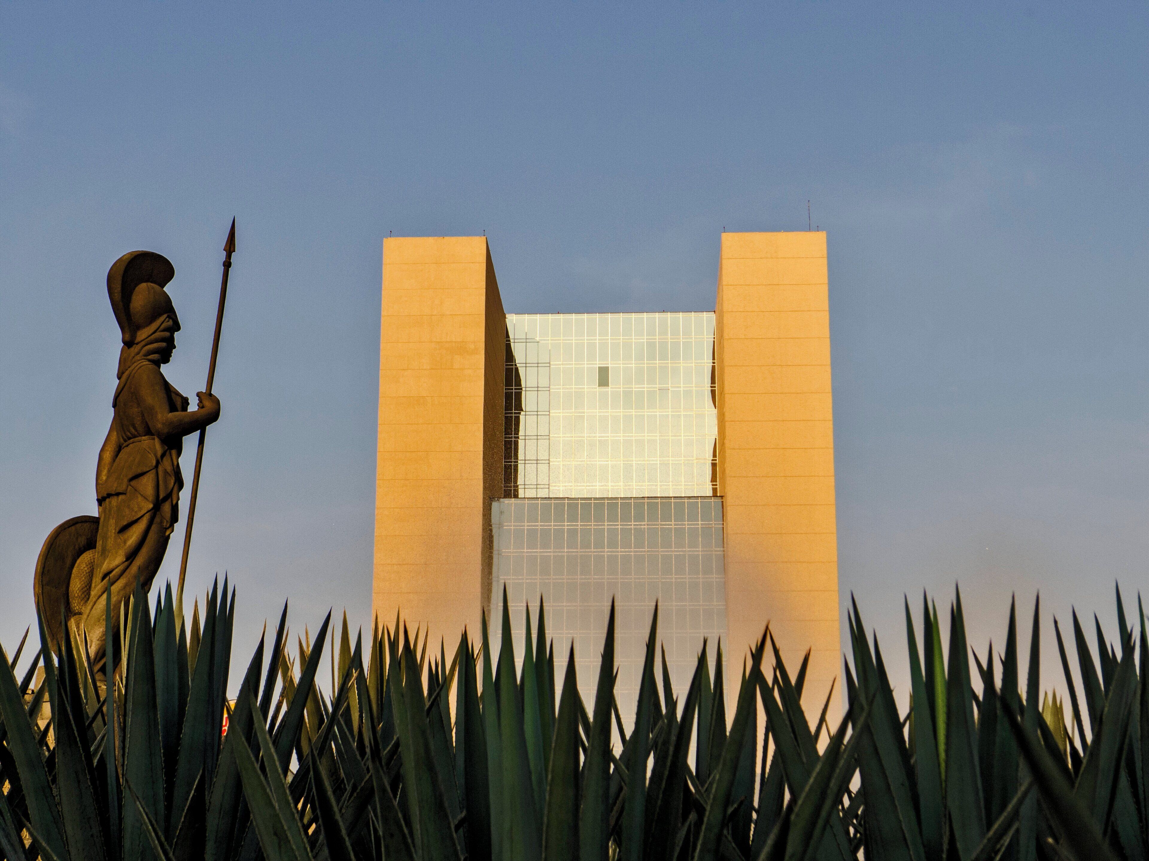 A statue of a figure holding a spear, a modern building with a glass facade, and tall, spiky plants in the foreground.