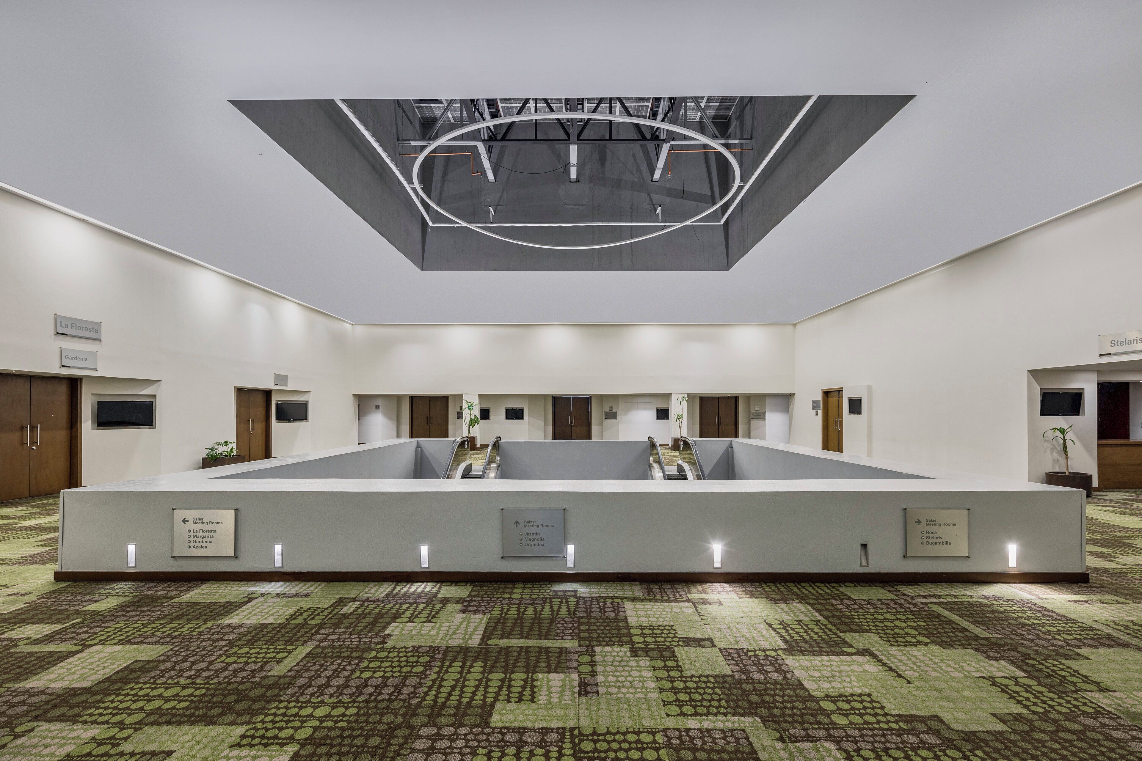 Un vestíbulo de hotel con una zona de estar central, un techo con diseño geométrico y señalización direccional para salas de reuniones.