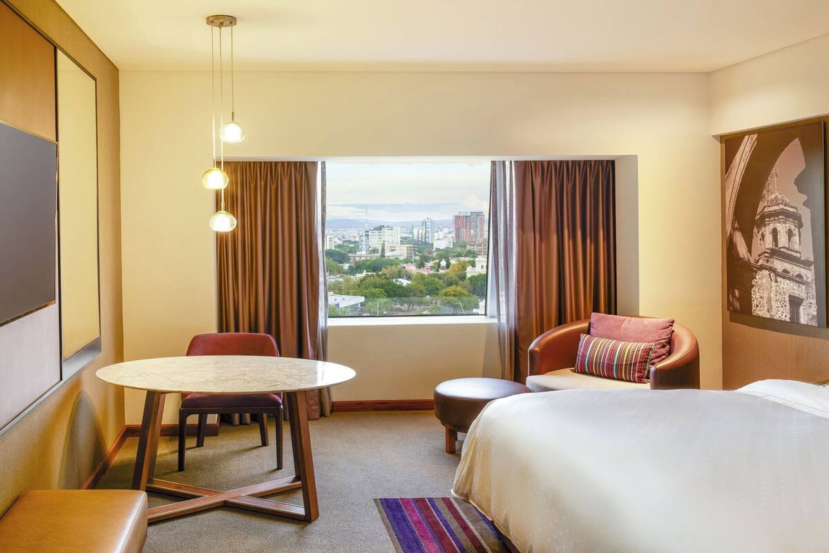Een hotelkamer met een bed, een bureau, een stoel, een televisie en uitzicht op een stadsbeeld.