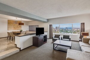 Habitación de hotel moderna con zona de comedor, televisor de pantalla plana, sofá y un amplio ventanal con vistas a la ciudad.
