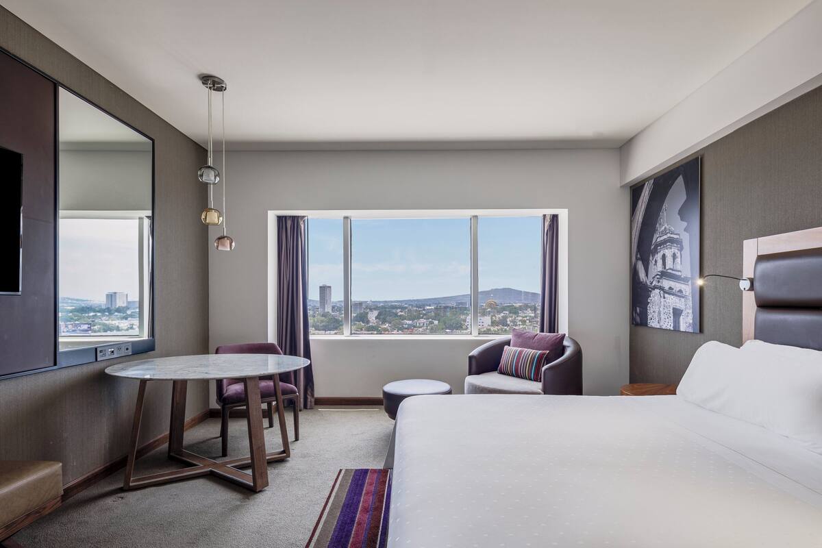 Hotelkamer met een bed, een bureau, een stoel, een kleine ronde tafel en uitzicht op de stadsgezicht door grote ramen.