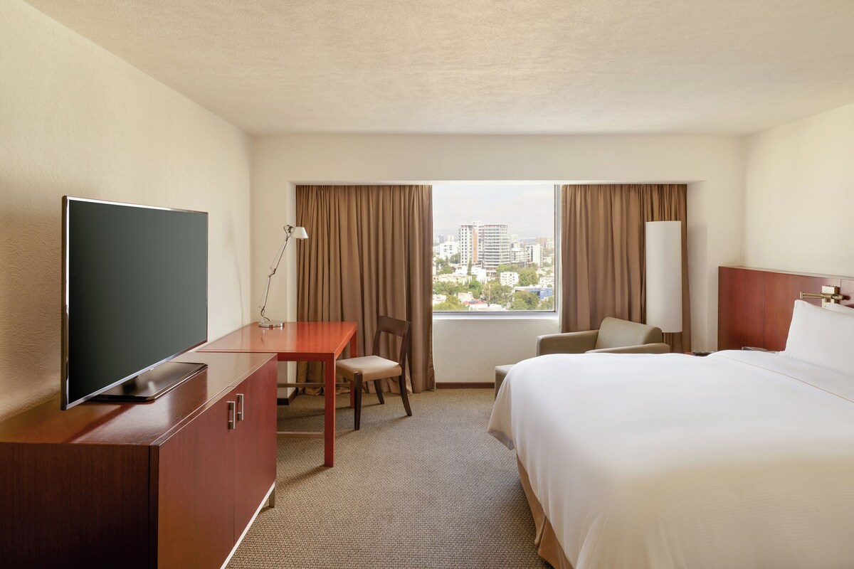 A hotel room with a bed, a desk with a TV, a chair, and a view of the city through the window.