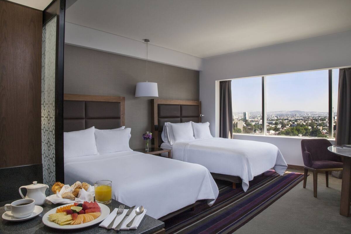 Een hotelkamer met twee bedden, een ontbijtgedeelte, een raam met uitzicht op de stad en een klein tafeltje met een stoel.