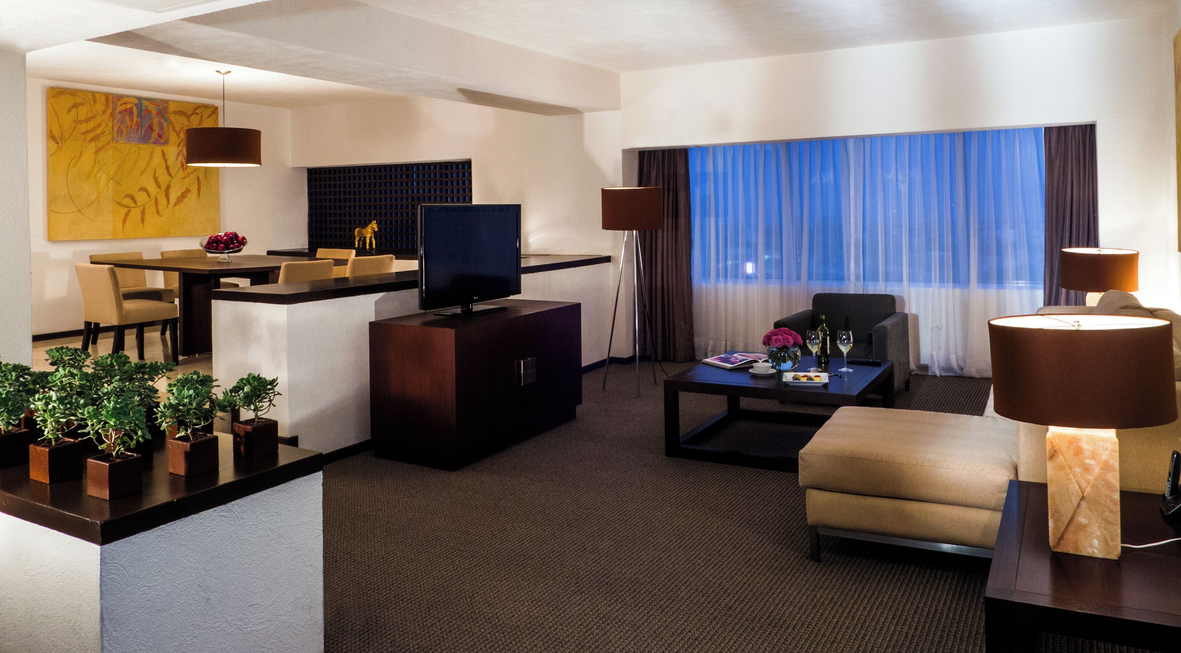 A modern hotel room with a dining area, a flat-screen TV, a sofa, a coffee table, and a lamp.