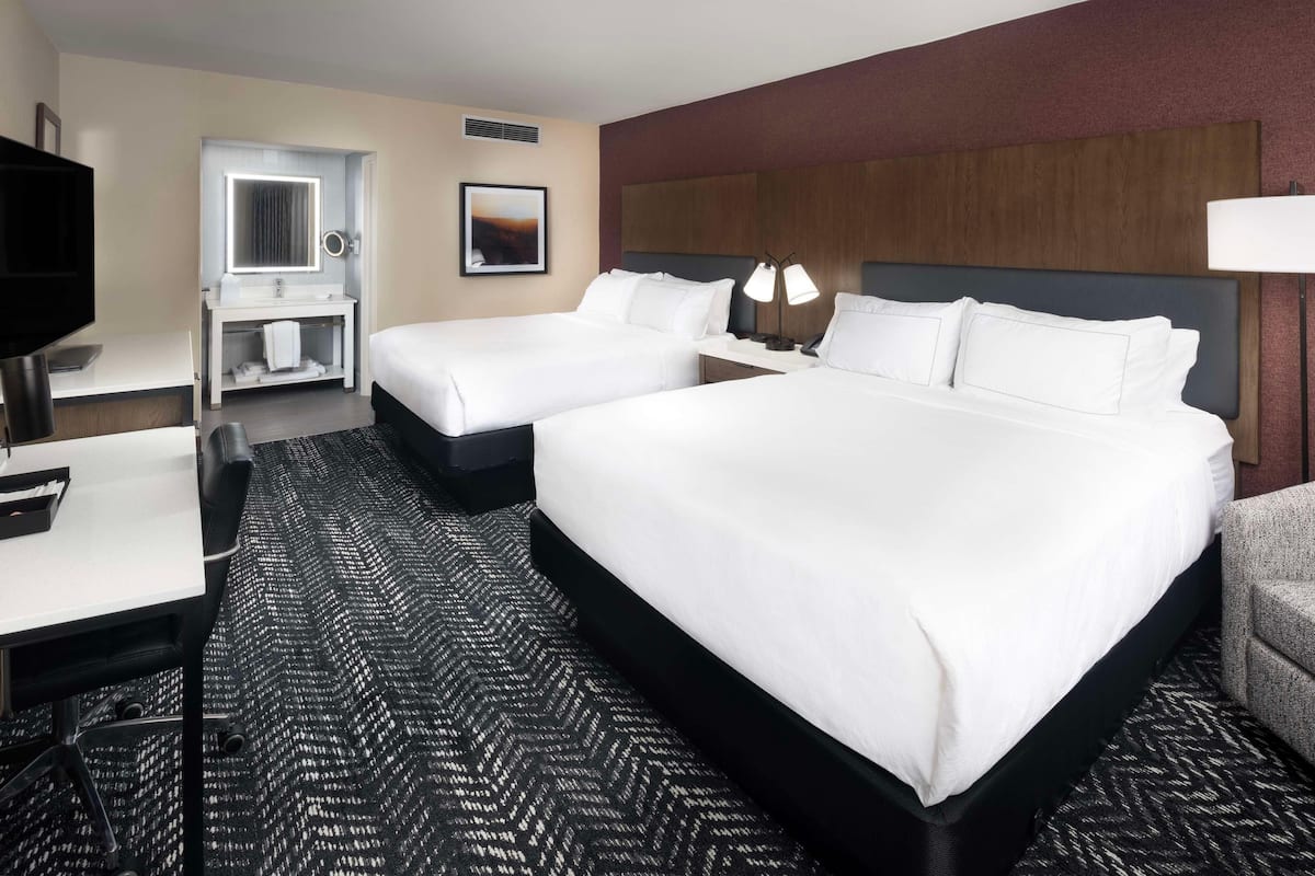 A hotel room with two beds, a desk, a vanity with a mirror, and a TV.