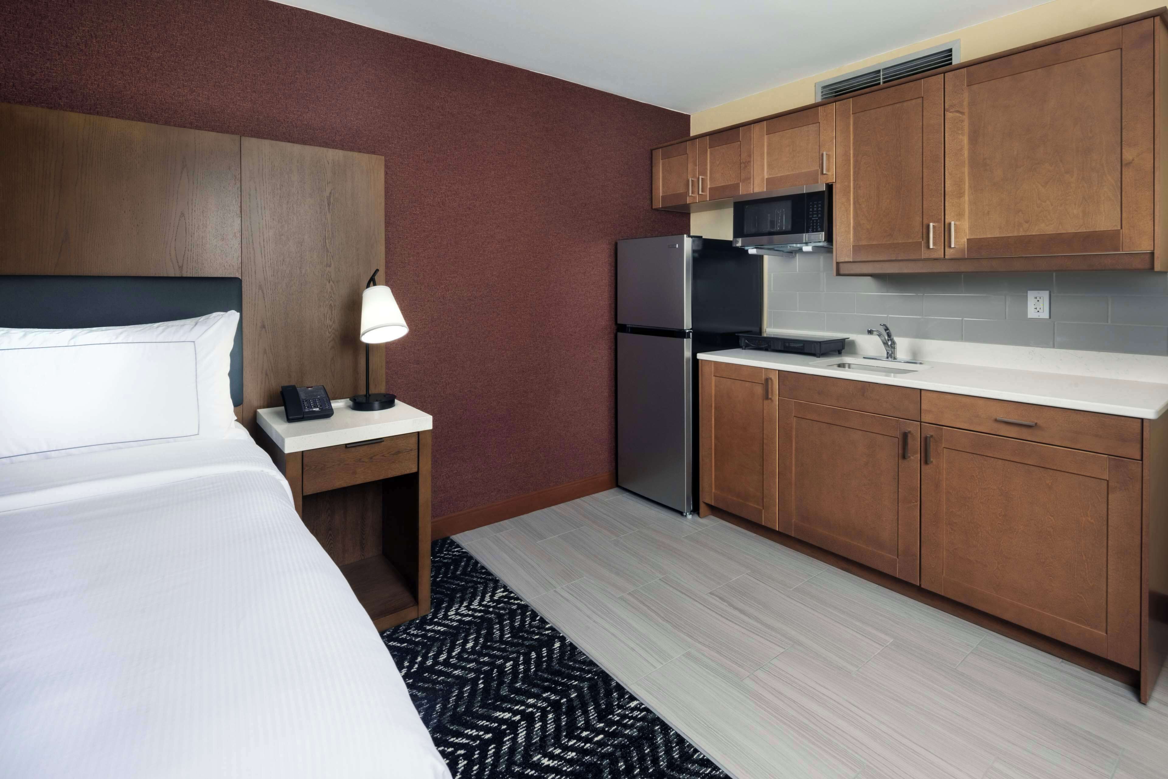 Quarto de hotel compacto com cama, mesa de cabeceira, luminária, cozinha compacta com armários de madeira e micro-ondas.