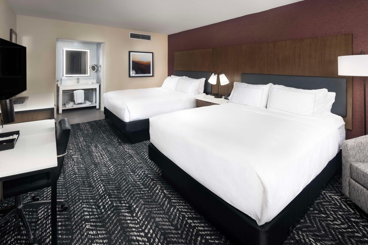 A hotel room with two beds, a desk, a vanity with a mirror, and a TV.