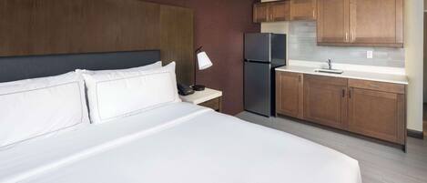 A hotel room with a large bed, a nightstand, a refrigerator, wooden cabinets, and a small kitchen area.