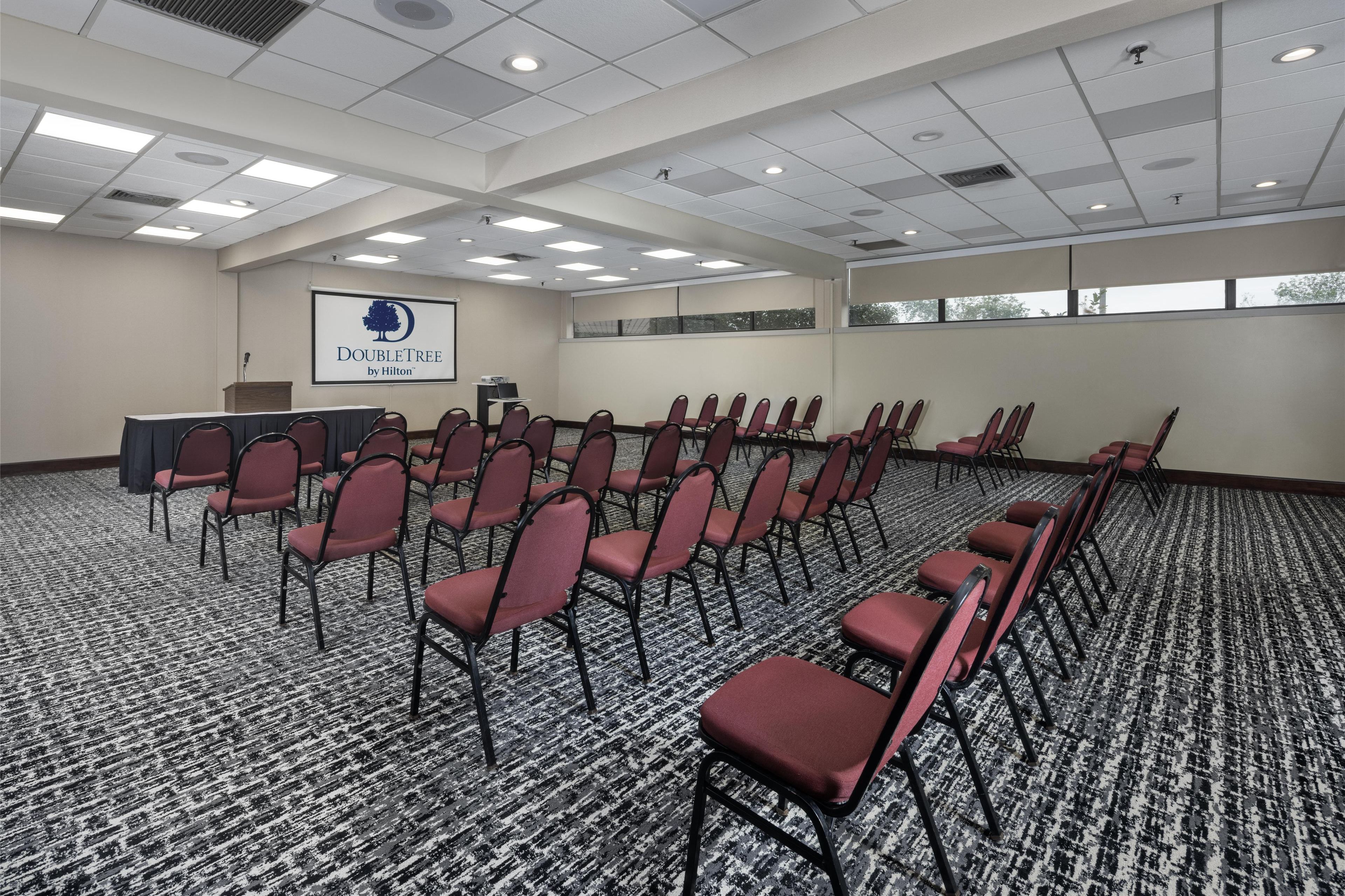 A conference room with rows of red chairs, a podium, and a screen displaying the DoubleTree by Hilton logo.