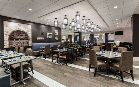 A modern hotel dining area with multiple tables and chairs, a large arched fireplace, and hanging pendant lights.