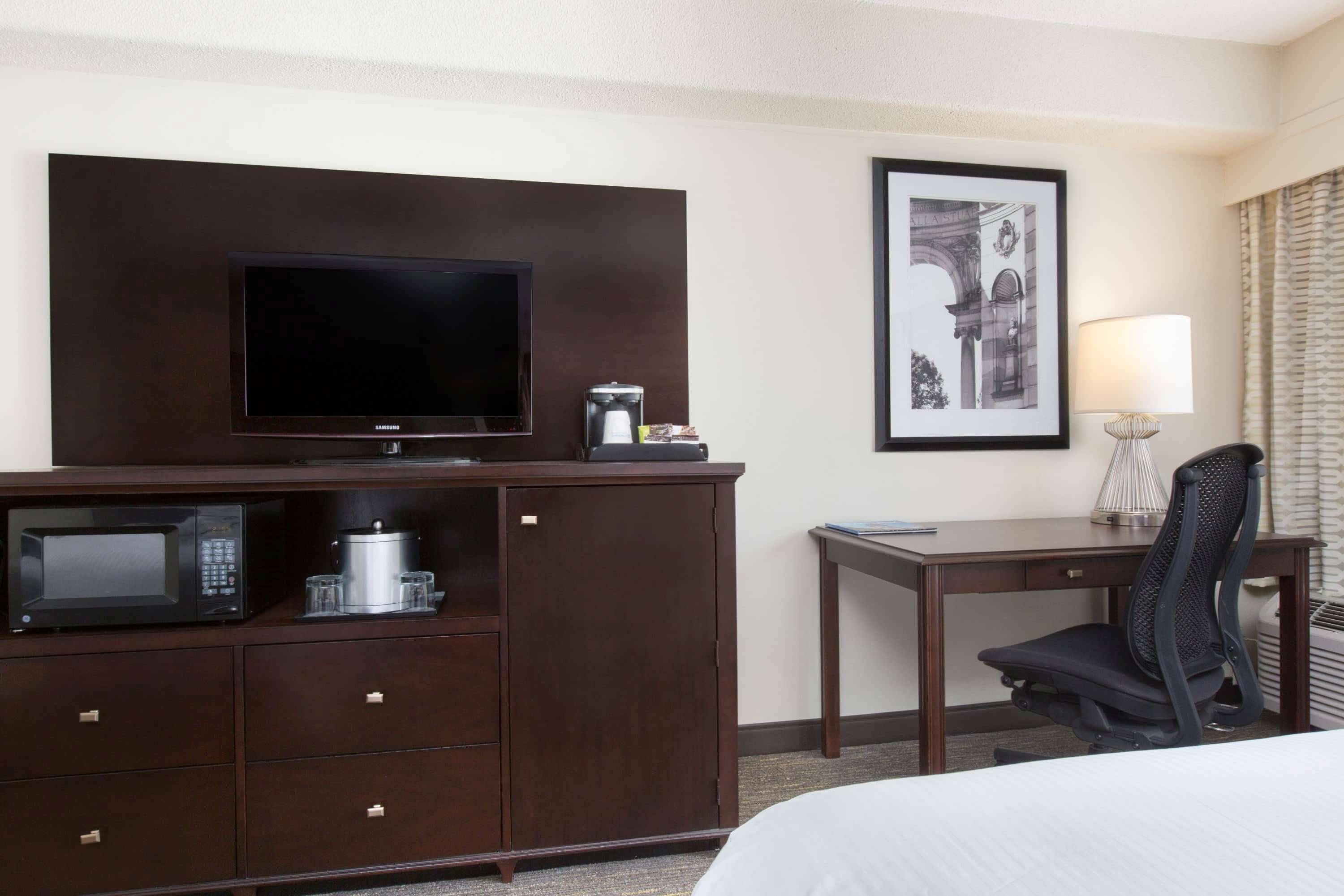 Habitación de hotel con televisor de pantalla plana, microondas, cafetera, escritorio con silla y un cuadro en la pared.