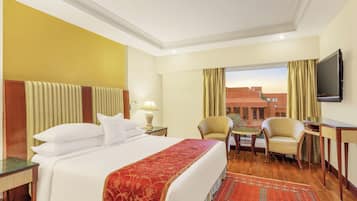 Une chambre d’hôtel avec un lit, deux chaises, une petite table, un téléviseur et une fenêtre avec des rideaux.