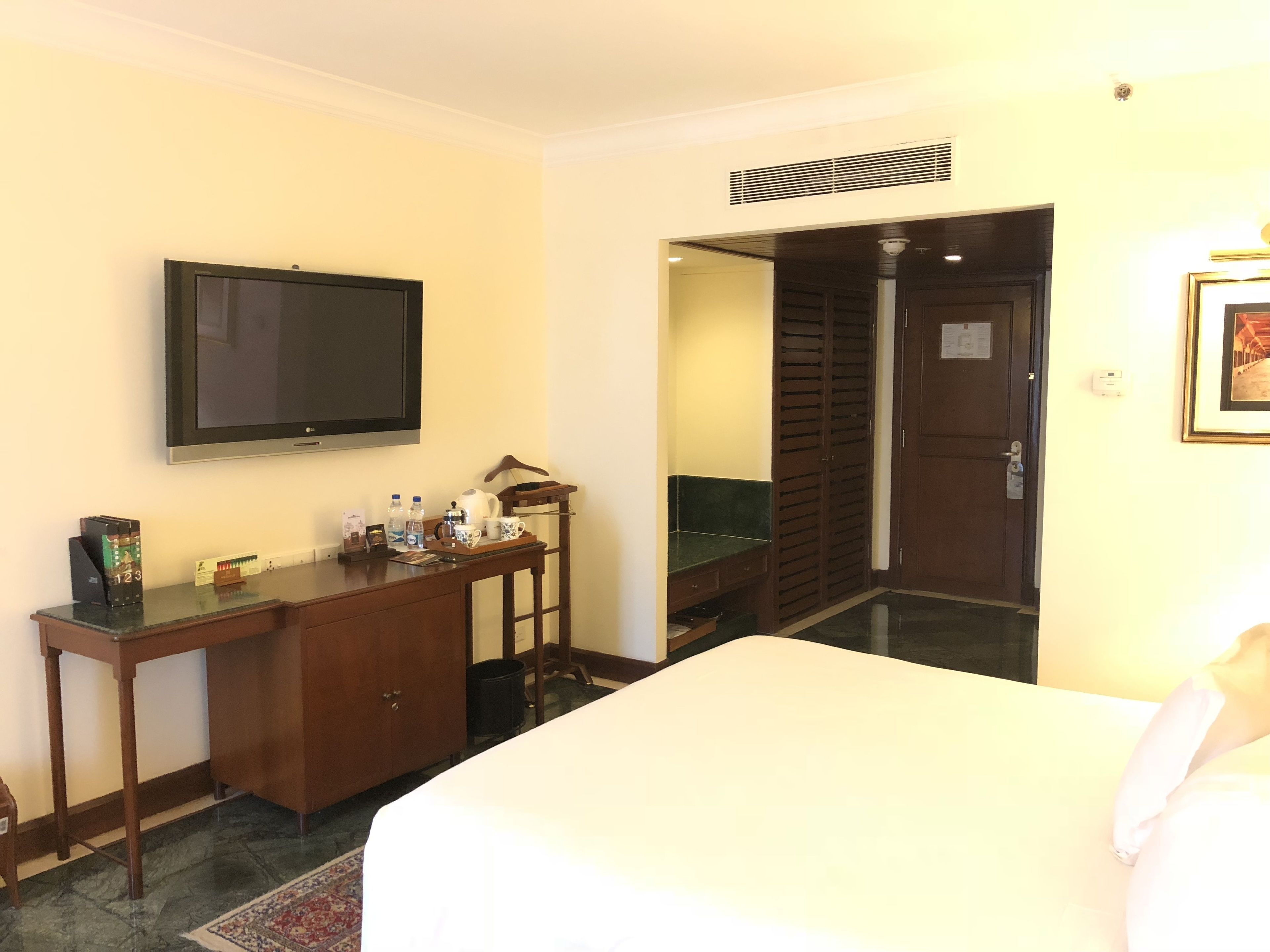 A hotel room with a bed, a desk with a flat-screen TV, a mirror, a wardrobe, and a door leading to another room.