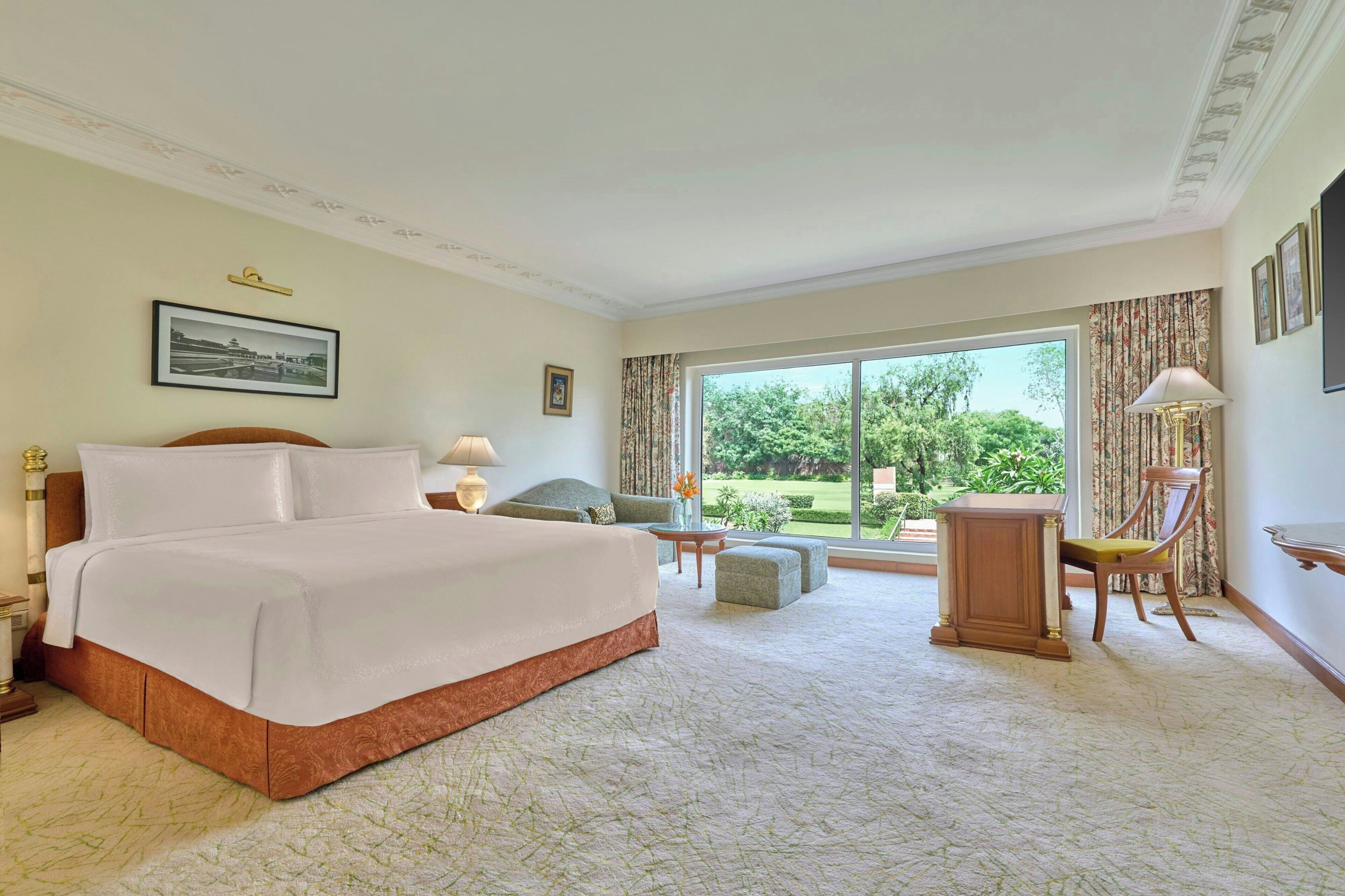 Habitación de hotel con una cama grande, un área de descanso con una mesa y sillas, y vista a un jardín por la ventana.
