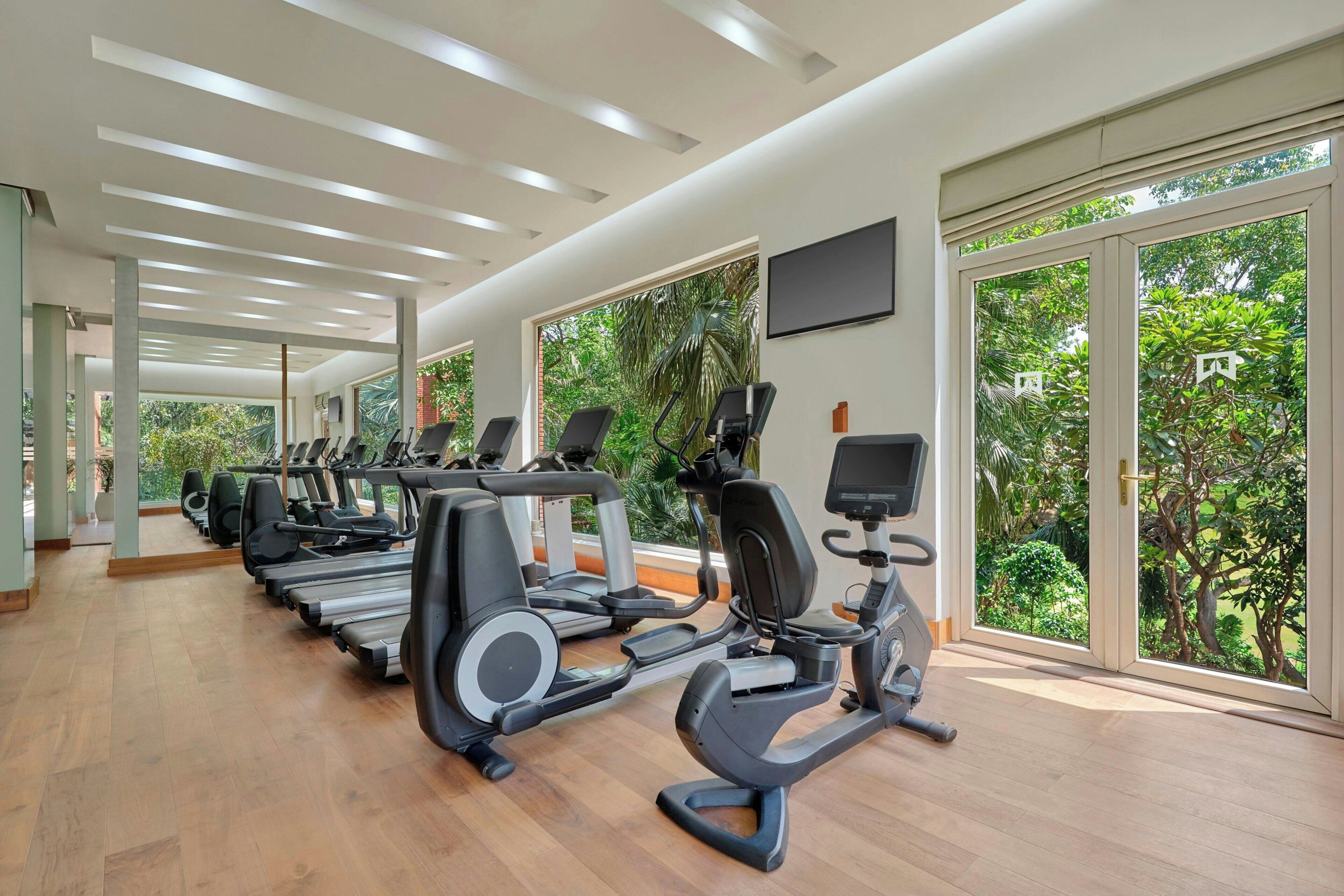 Une salle de sport équipée de plusieurs tapis de course et de vélos d’exercice, de grandes fenêtres offrant une vue sur la verdure et d’un plafond au design moderne.