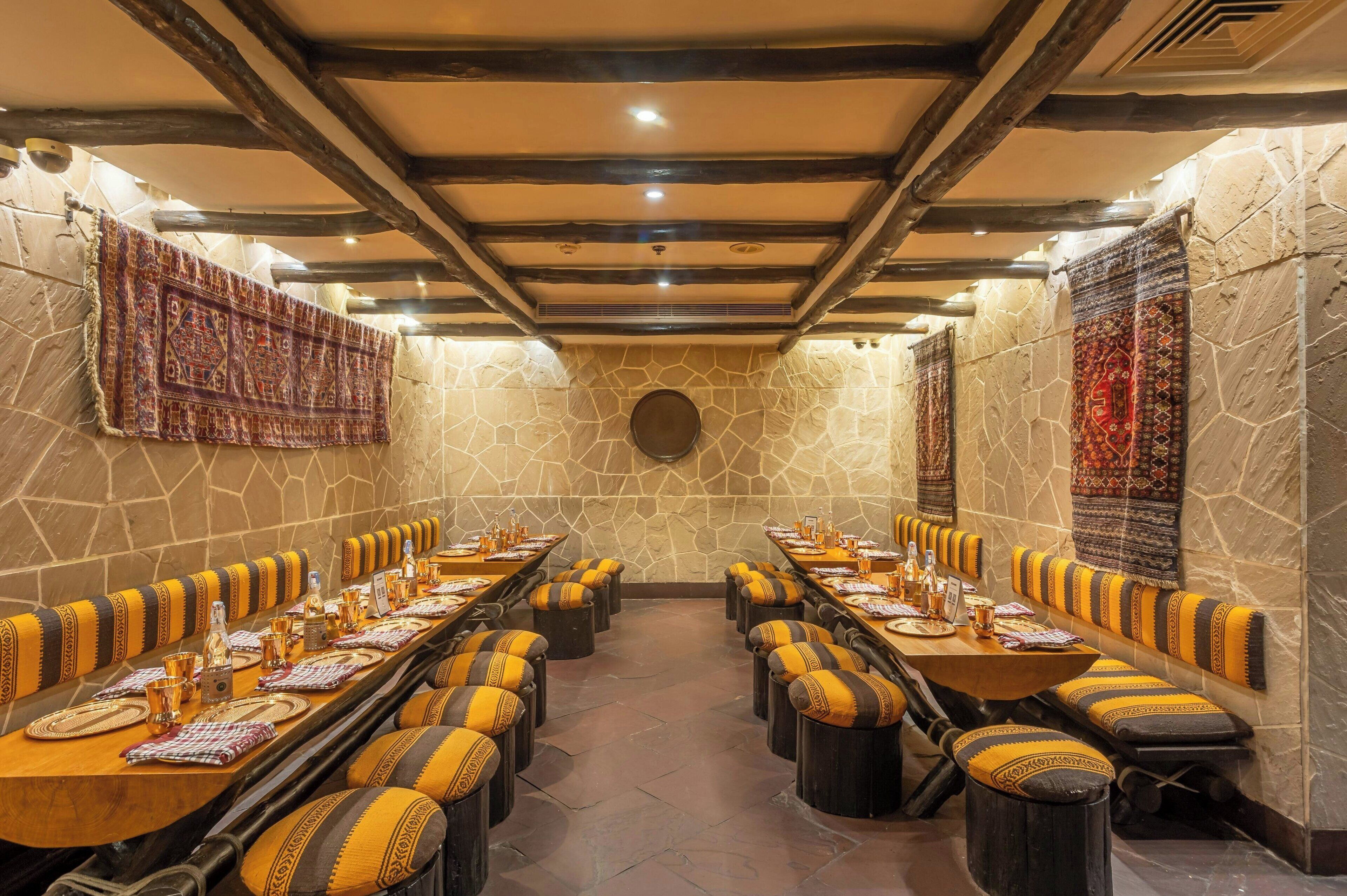 A long dining table with multiple chairs, each with a patterned cushion. The room has stone walls and a wooden ceiling with exposed beams.