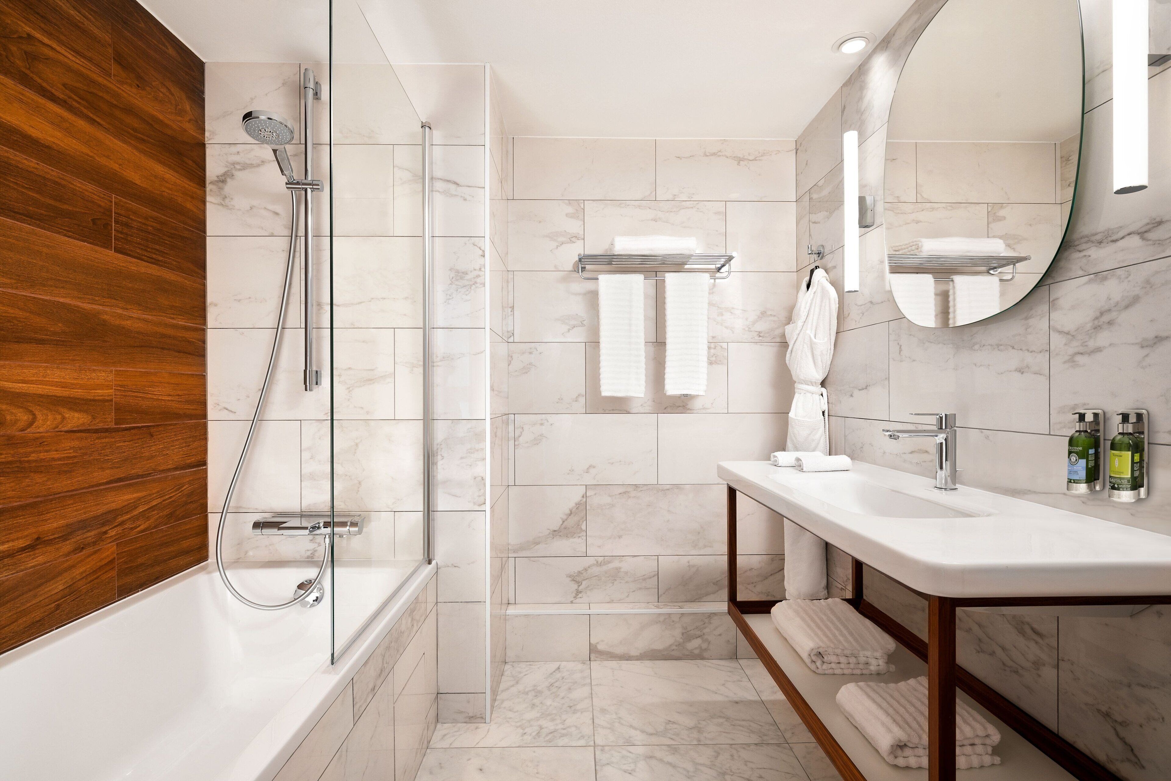 Une salle de bain moderne avec une douche à parois de verre, un mur d’accent en bois, un sol carrelé en marbre, un lavabo blanc et un grand miroir.