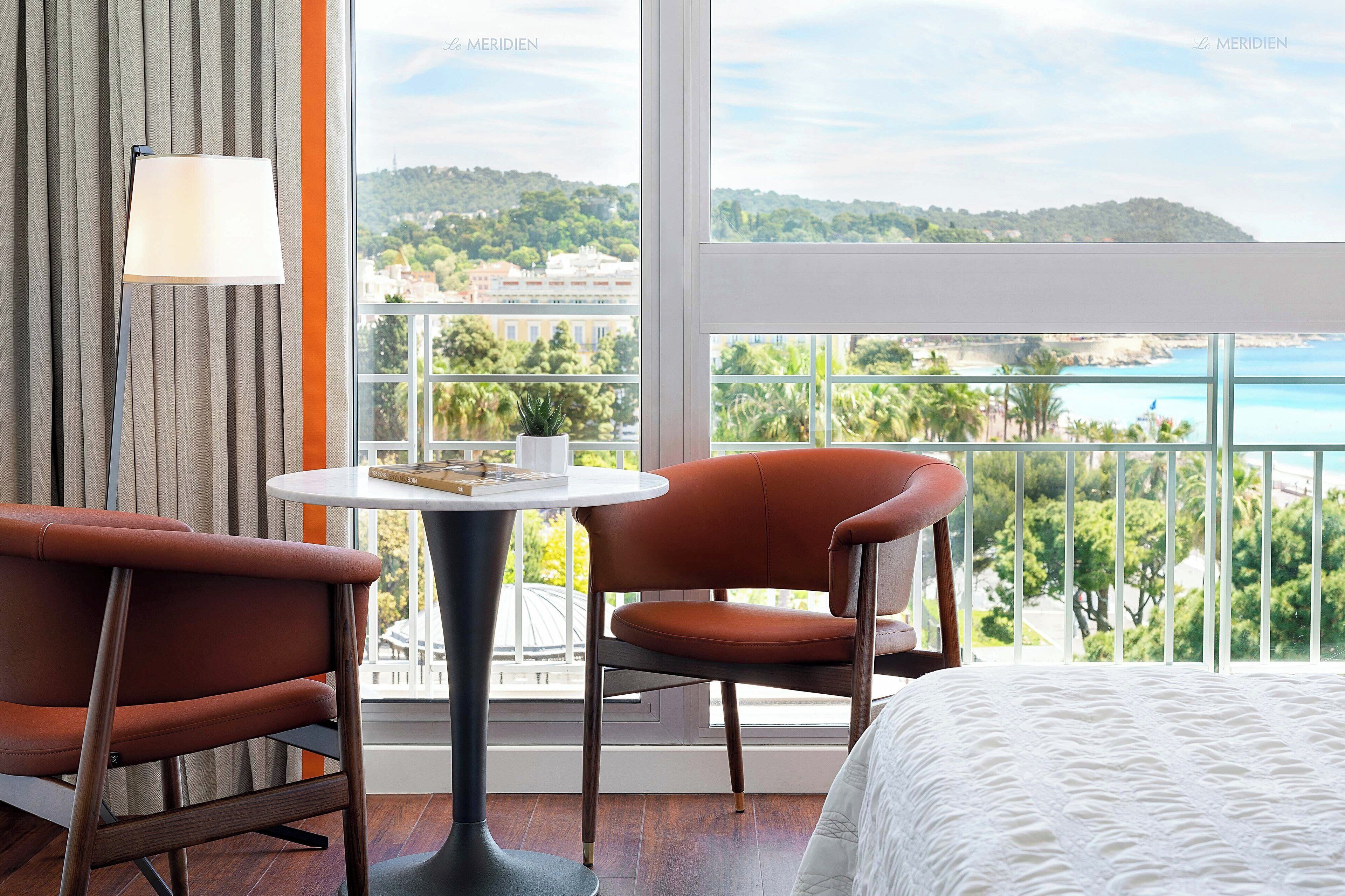 Habitación de hotel con cama, dos sillas, una mesita y vistas a un paisaje costero.