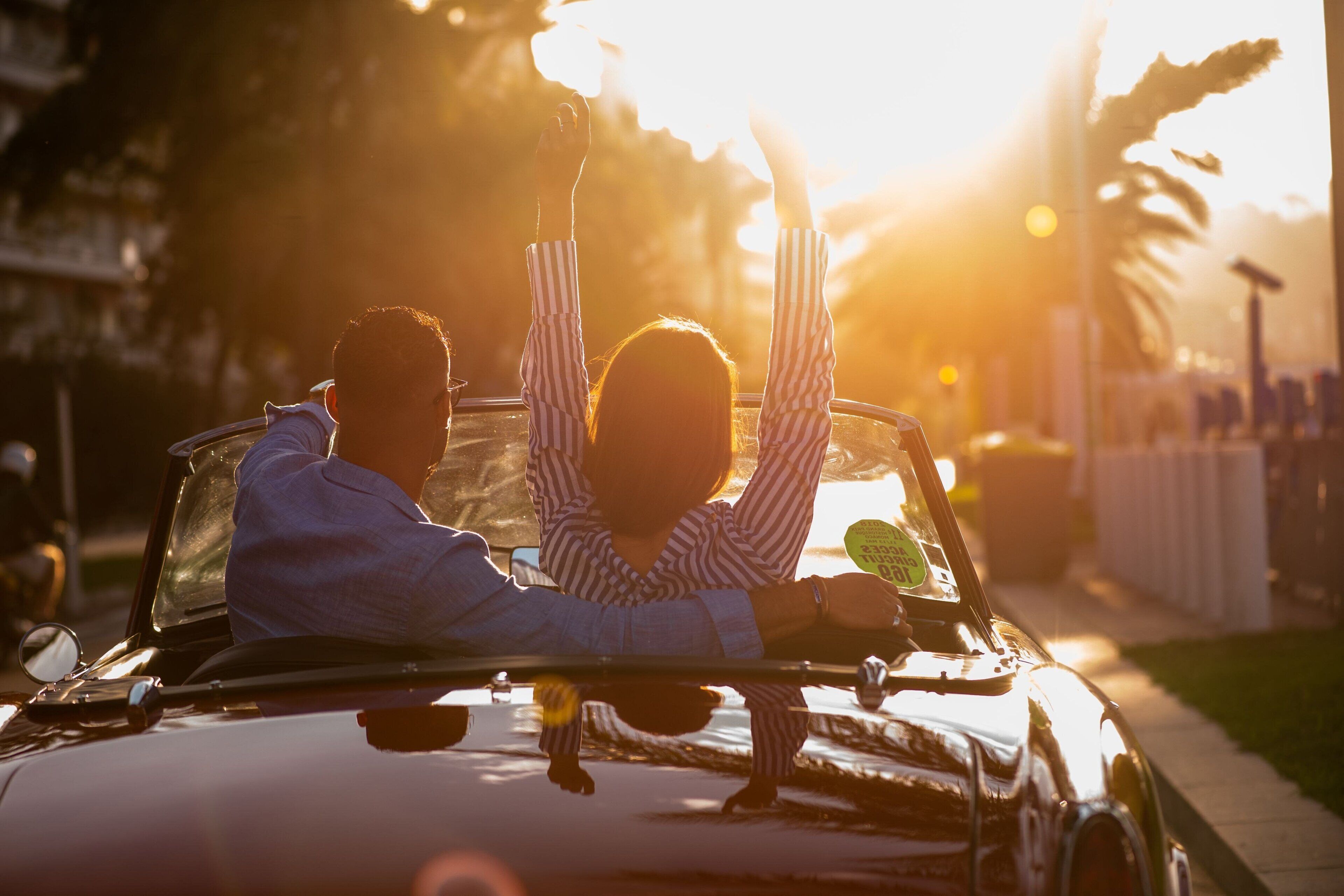 A convertible car with two people inside, one raising their hand.