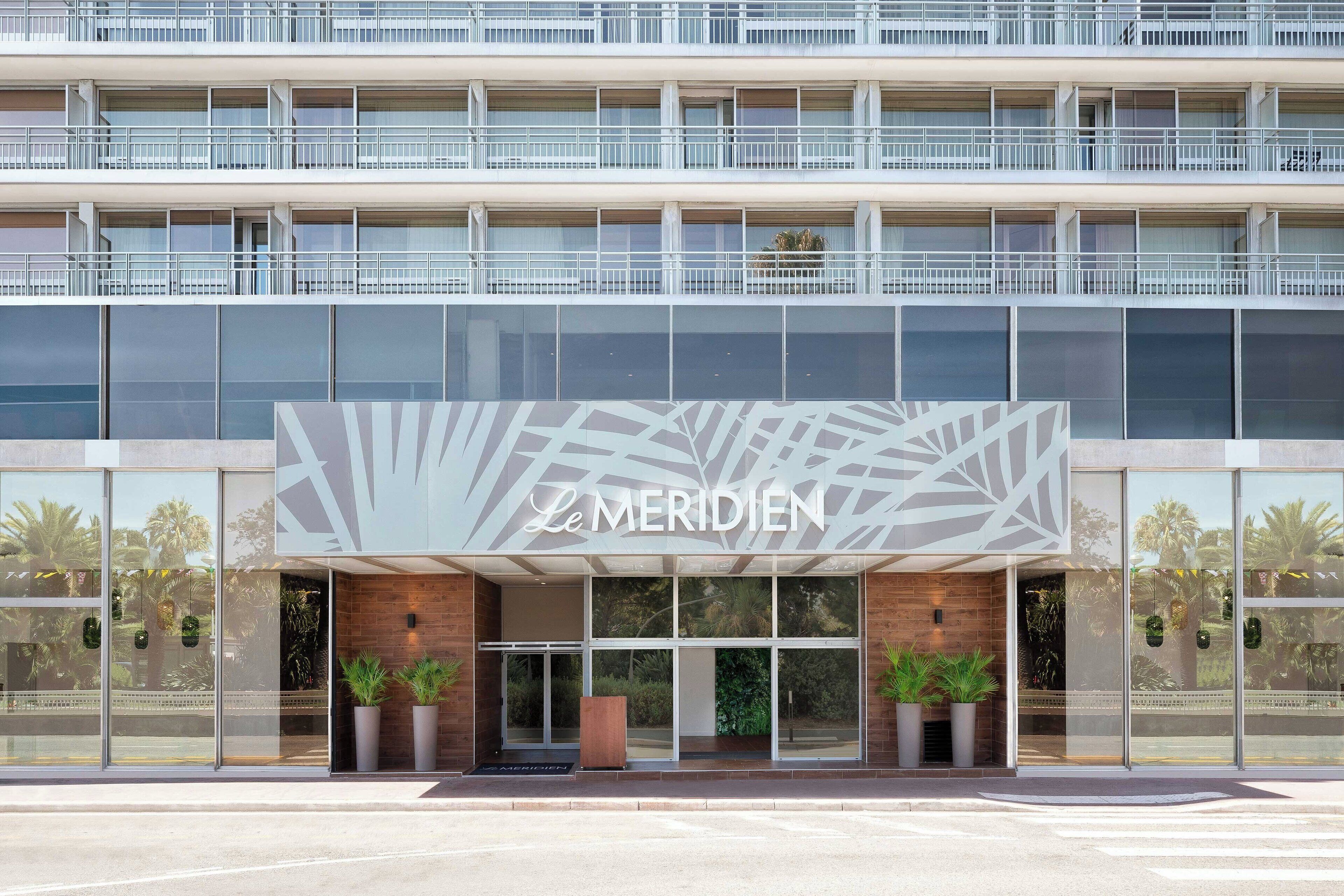 A hotel entrance with a modern facade, large glass windows, and a prominent sign reading "Le Meridien".