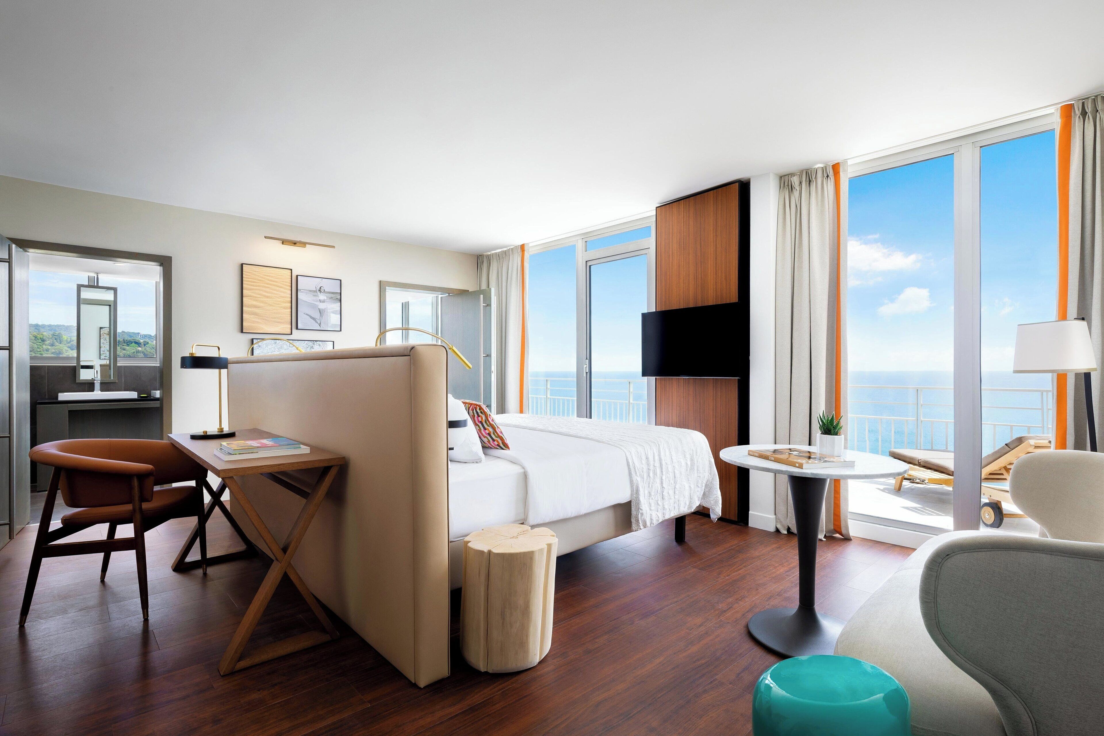 A modern hotel room with a large bed, a desk, a chair, a small table, and a TV.