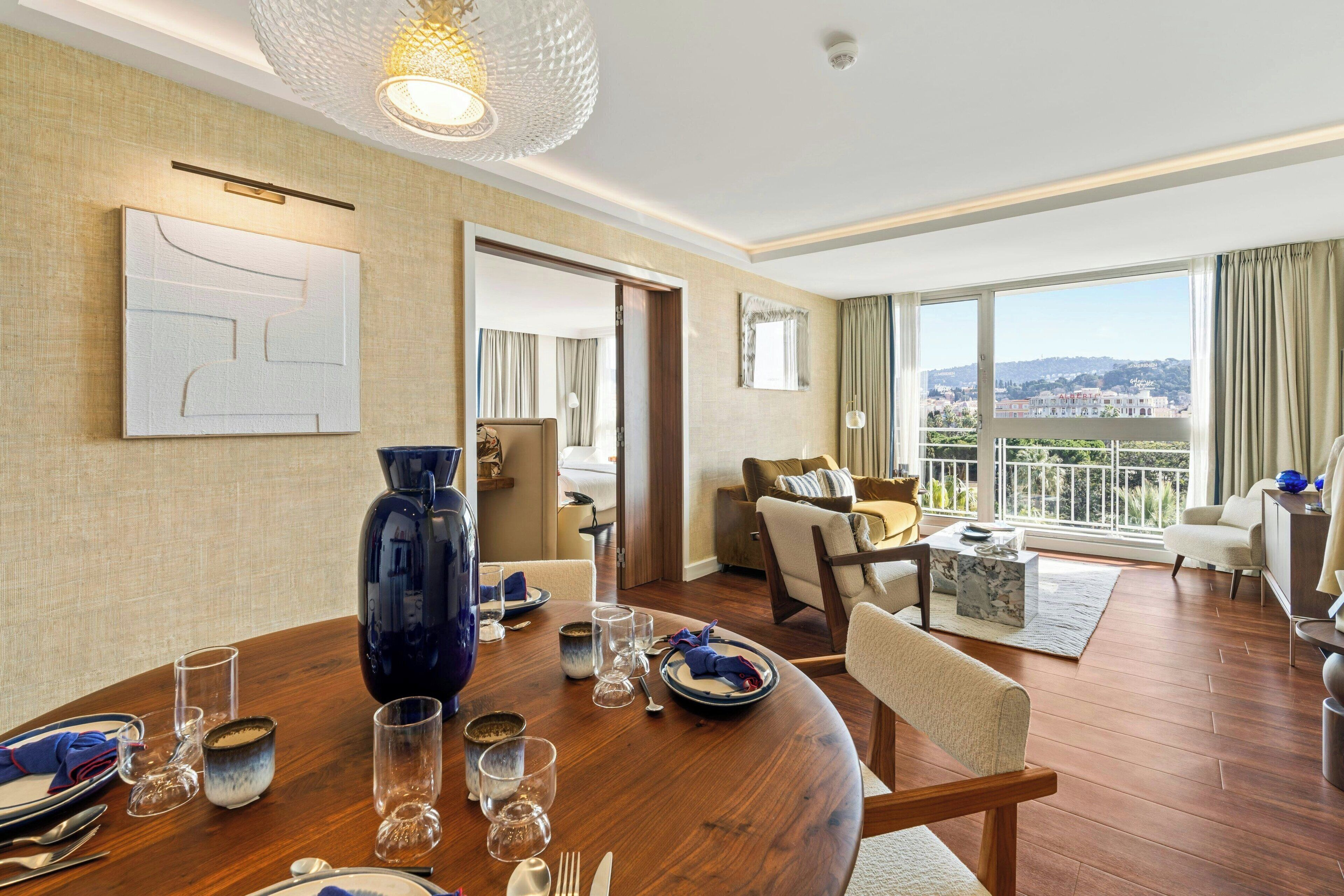 Une chambre d’hôtel moderne comprenant un coin repas, un salon avec un canapé et des fauteuils, une grande fenêtre donnant sur la ville et un vase décoratif posé sur la table du coin repas.