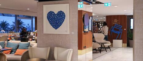 A modern hotel lobby with seating areas, a large heart-shaped artwork on the wall, and directional signs for amenities.