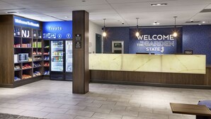 A hotel reception area with a marble counter, a 'Welcome to Garden State' sign, a 'Treats' section with snacks and beverages, and a 'NJ' sign indicating the location. - Hampton Inn Newark Airport (Newark)