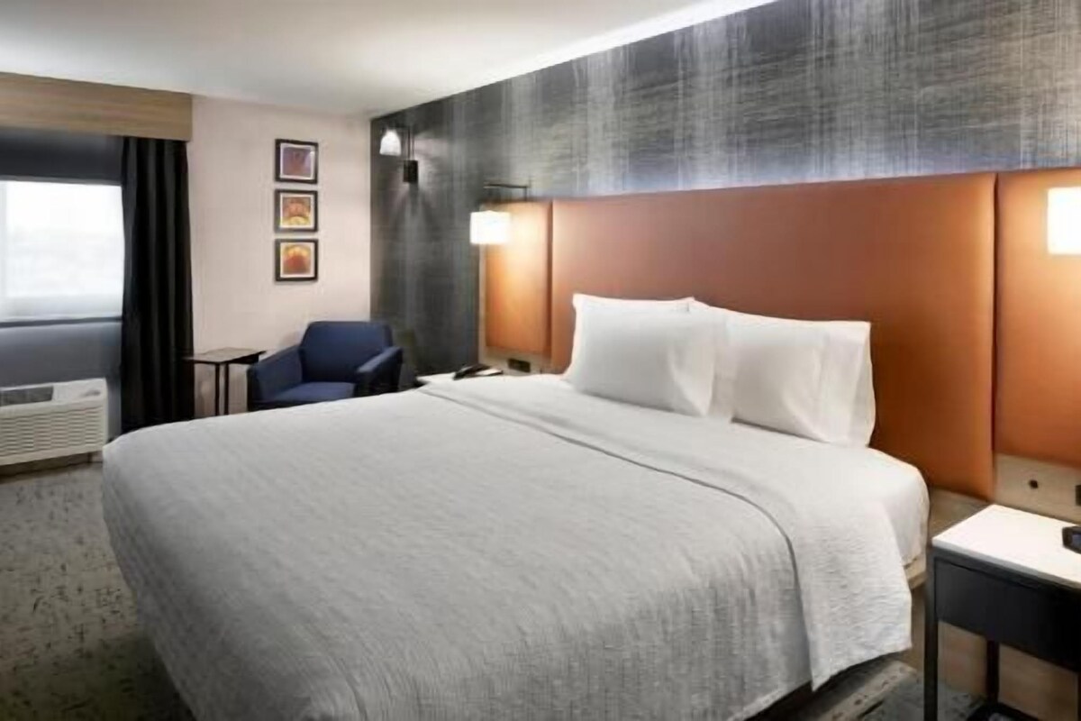 A hotel room with a large bed, a desk, a chair, and a wall-mounted lamp.